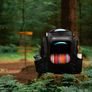 A black Discmania disc golf backpack filled with colorful discs, set on a mossy rock in a forest with a disc golf basket in the background.